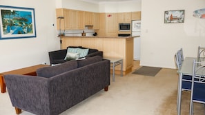 A living room with a sofa, a dining table with chairs, a kitchen area with cabinets and appliances, and a painting on the wall. - Oceania Cottage- Stone Real Estate Yamba (Yamba)