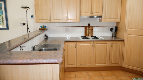 A kitchen with wooden cabinets, a stainless steel sink, a stove, and a tiled backsplash. - Oceania Cottage- Stone Real Estate Yamba (Yamba)