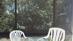 A view through a window with two white chairs and a table, with dense foliage and red flowers in the background. - Oceania Cottage- Stone Real Estate Yamba (Yamba)