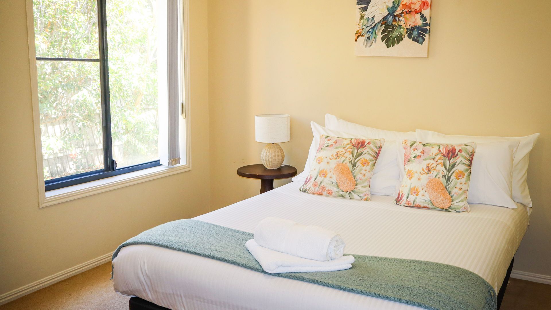 A neatly made bed with a white comforter and two floral pillows, a small round table with a lamp, and a window with a view of greenery.