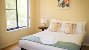 A neatly made bed with a white comforter and two floral pillows, a small round table with a lamp, and a window with a view of greenery. - Oceania Cottage- Stone Real Estate Yamba (Yamba)