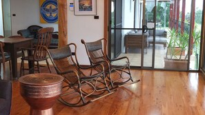 Una habitación con piso de madera, una mesa de comedor con sillas, dos mecedoras, una mesa de centro y un ventanal corredero que da a un espacio exterior con vista a follaje.