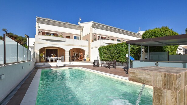 A swimming pool with a wooden deck, a covered seating area, and a building with arched doorways in the background.