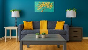 A living room with a teal wall, a gray sofa with yellow cushions, a coffee table, two side tables with lamps, and a painting of a tree. - Dreamy Group-Friendly 2BR | 2Kings w/ BQQ,Gym+Pool (Houston)