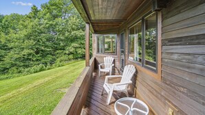 Um deck de madeira com duas cadeiras Adirondack brancas e uma mesa de vidro, com vista para um gramado bem cuidado e árvores densas.