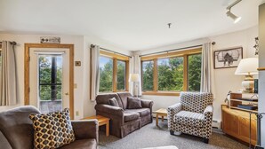 Uma sala de estar acolhedora com dois sofás, uma poltrona estampada, uma mesa pequena e um abajur. Janelas amplas proporcionam vista para o verde lá fora.