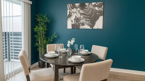 Un coin repas avec une table ronde dressée pour quatre personnes, entourée de chaises de couleur beige. Un grand tableau abstrait est accroché au mur de couleur turquoise, au-dessus de la table.