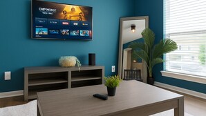 Un salon aux murs de couleur turquoise, avec un téléviseur fixé au mur diffusant un film, une table basse en bois, une plante en pot et un grand miroir qui reflète la pièce.