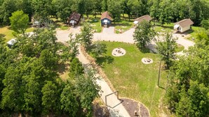 Una vista aerea di un campeggio con diverse cabine, un'area comune con posti a sedere disposti a cerchio, e un vialetto lastricato che collega le cabine.