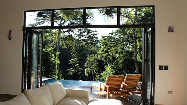 A modern interior with a sofa and wooden chairs near a glass door leading to an outdoor area with a pool and lush greenery. - Chial Reserve (San Ignacio)
