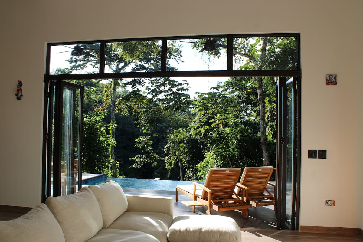 Un interior moderno con un sofá y sillas de madera cerca de un ventanal que da a un área exterior con piscina y abundante vegetación.