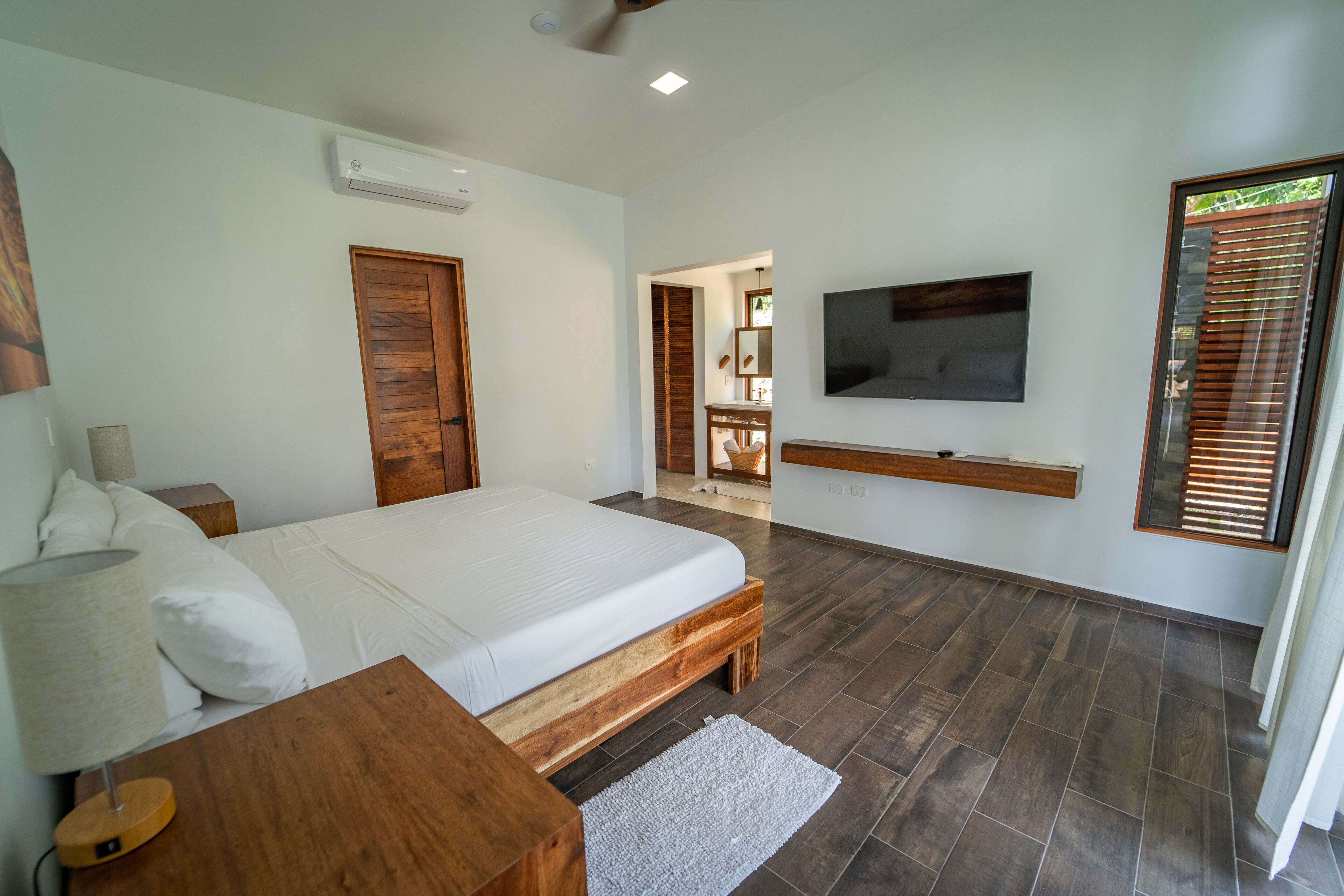 Une chambre à coucher avec un lit, un téléviseur fixé au mur, une armoire en bois et une fenêtre avec des stores.