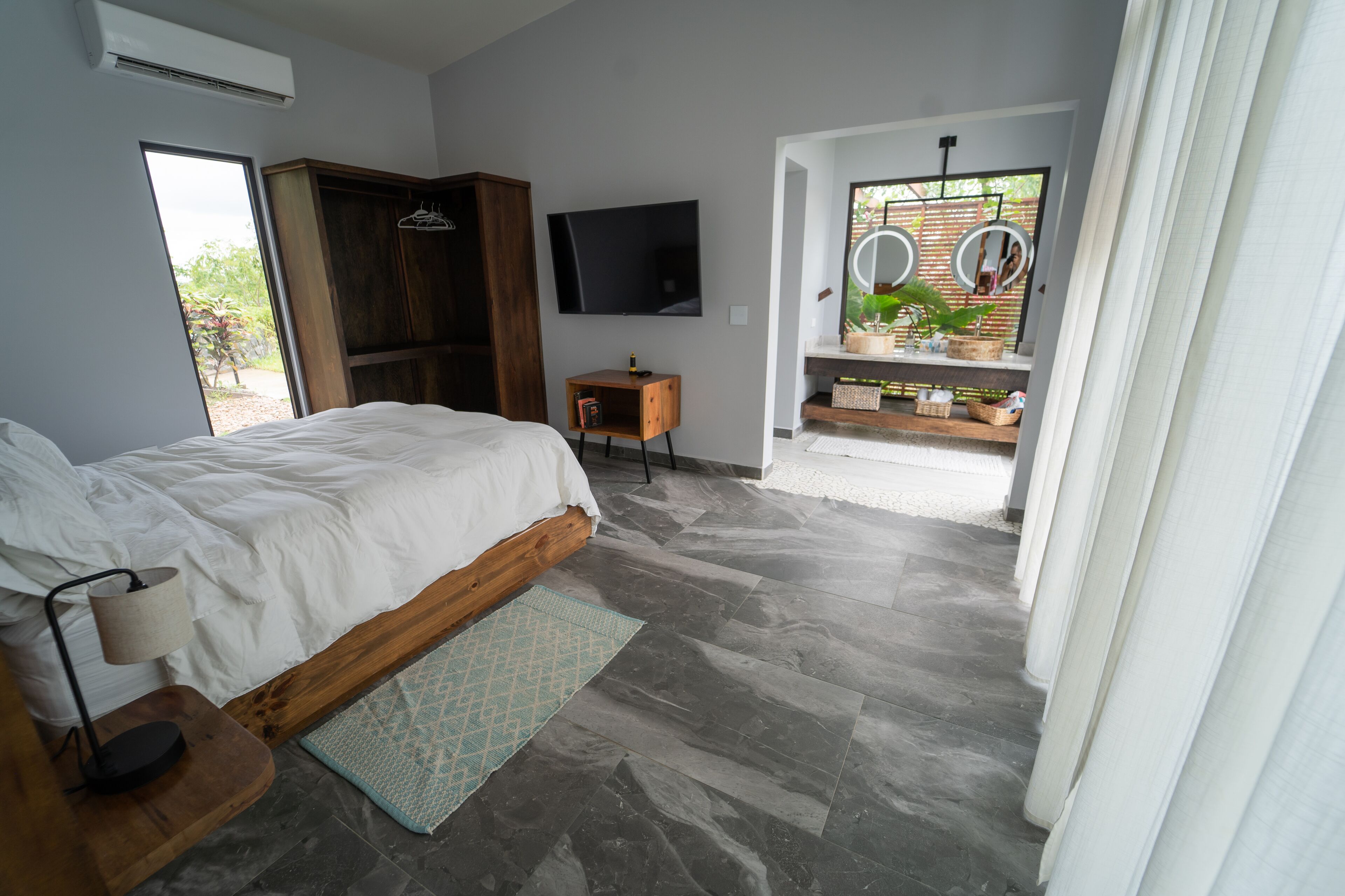 A bedroom with a bed, a TV, a wardrobe, a small table with a lamp, and a bathroom with a mirror and plants.