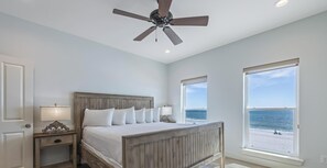 A bedroom with a wooden bed, two bedside tables, a ceiling fan, and large windows overlooking the beach.