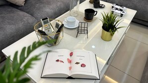 Un salon moderne avec une table basse en verre, un livre, une plante en pot, une machine à café et des manettes de jeu.