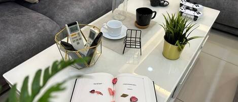 A modern living room with a glass coffee table, a book, a potted plant, a coffee maker, and gaming controllers.