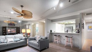 A modern living room with a grey sofa, a smaller grey armchair, and a large ceiling fan. There is a kitchen area with a white countertop and a mirror above it.