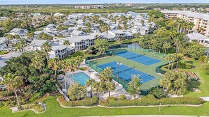 An aerial view of a residential area with multiple houses, a tennis court, and a swimming pool surrounded by palm trees and greenery. - Gorgeous bay views from this ground floor Siesta Key Condo - GBB 1360 (Siesta Key)