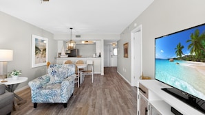 A modern living room with a flat-screen TV, a sofa, a dining area, and a kitchen in the background. - Gorgeous bay views from this ground floor Siesta Key Condo - GBB 1360 (Siesta Key)