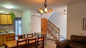 A dining area with a wooden table and chairs, a brown leather sofa, a staircase, and a kitchen with wooden cabinets and a hanging plant.