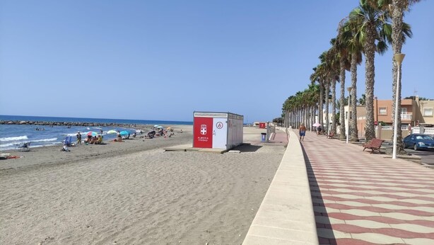 A beach with a red and white sign, palm trees, and a paved walkway.