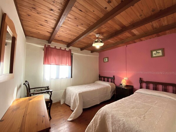 A bedroom with two beds, wooden ceiling beams, a window with a pink curtain, and a small table with a lamp.