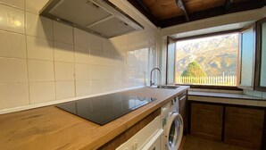 A kitchen with white tiles, a wooden countertop, a built-in stove, a washing machine, and a window with a view of a fence and trees. - Casa El Marizal 2 Arriondas (Montealea)