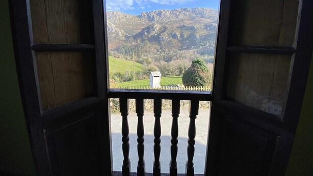 Vista a través de una ventana con marco de madera y barandilla, que da a un paisaje montañoso con un edificio pequeño y un área cercada.