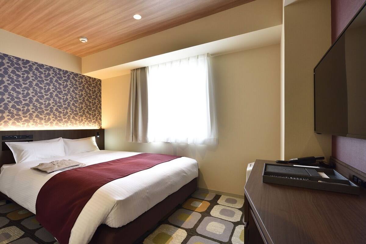 Una habitación de hotel con cama, escritorio, televisión, pared con diseño y ventana con cortinas.