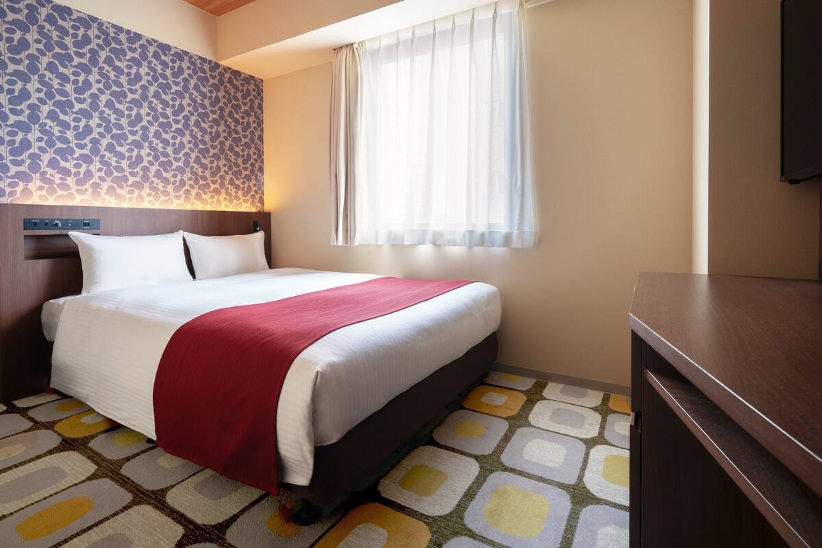 Habitación de hotel con cama, cabecera con panel de control, ventana con cortinas translúcidas y alfombra estampada.