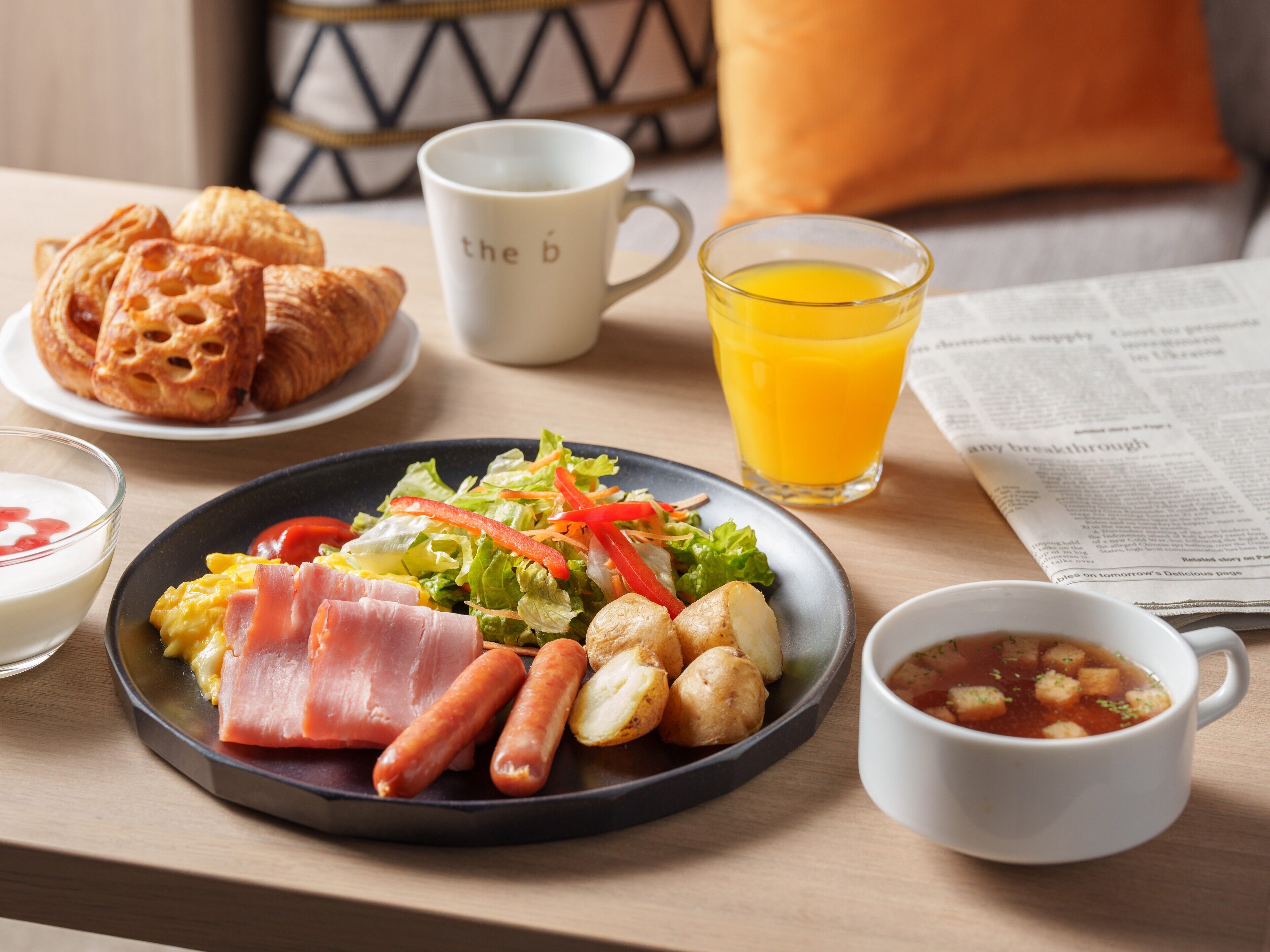 A breakfast setting with croissants, a plate of eggs, ham, sausages, and a salad, along with a glass of orange juice and a cup of coffee.
