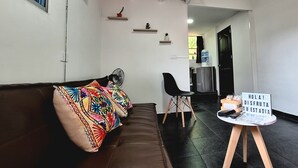Een woonkamer met een bruine bank, een kleurrijk kussen, een zwarte stoel en een kleine tafel met een bordje.
