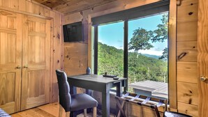 A wooden cabin room with a table and chair, a television, and a large window with a view of a forest.