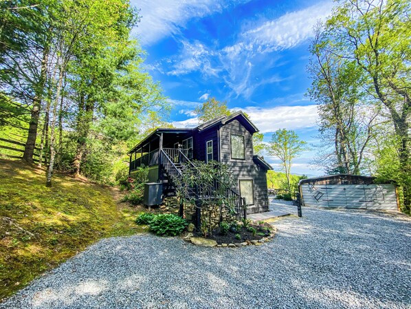 A rustic cabin with a porch, surrounded by trees and a gravel driveway.