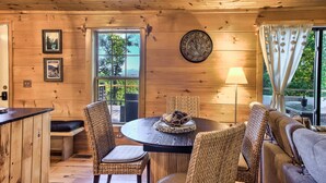 A wooden cabin interior with a round dining table, wicker chairs, a sofa, a wall clock, and a window with curtains.