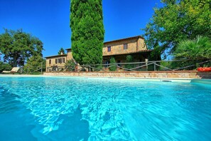 Une piscine à l’eau d’un bleu limpide, entourée d’un mur de pierre et d’une clôture en bois. Au fond, on aperçoit un bâtiment en pierre avec un toit en tuiles et un grand arbre.