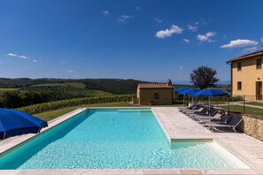 A swimming pool with lounge chairs and umbrellas, surrounded by a stone wall and a view of rolling hills.
