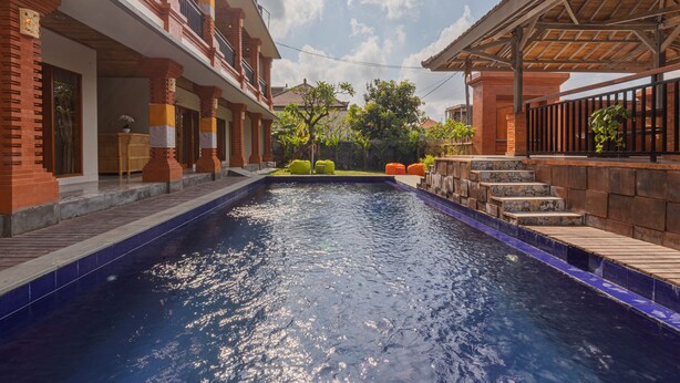 A swimming pool with a wooden deck and steps leading into the water, surrounded by a brick building with balconies and outdoor seating.
