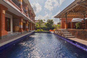 A swimming pool with a wooden deck and steps leading into the water, surrounded by a brick building with balconies and outdoor seating.