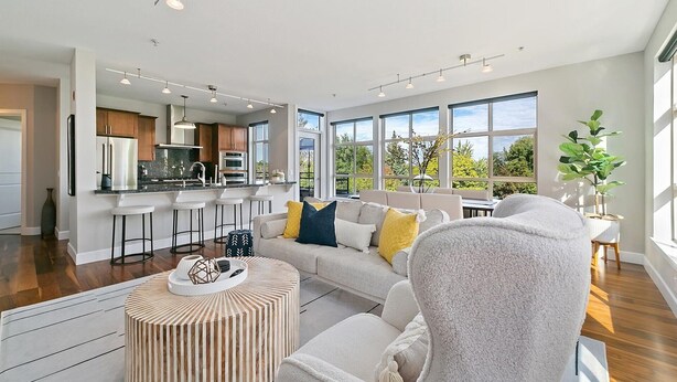 A modern living room with a sofa, ottoman, and a coffee table. There is a kitchen in the background with a refrigerator, oven, and bar stools.