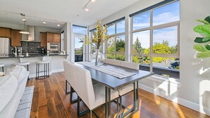 A modern kitchen with a dining area, large windows, and a view of greenery.