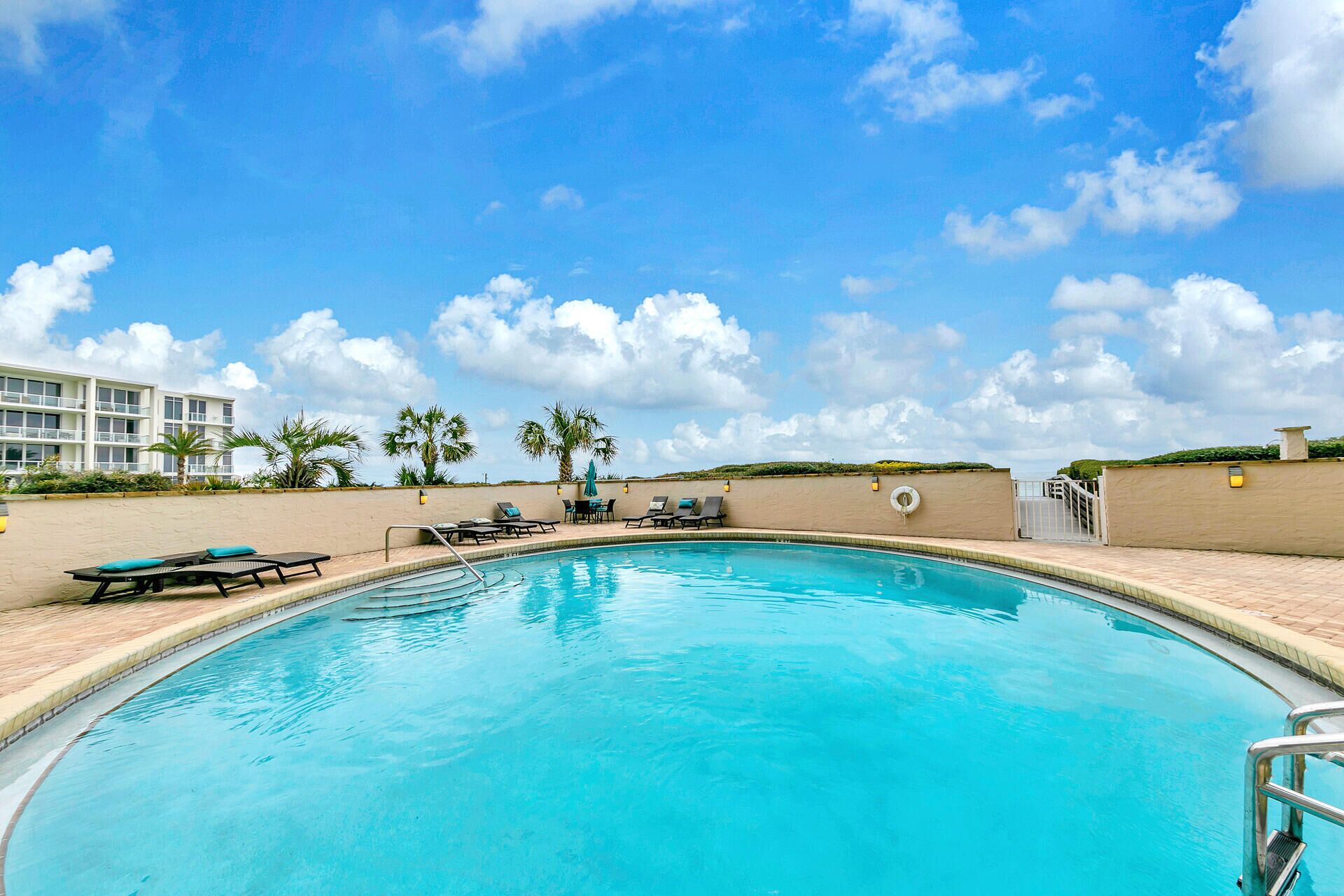 A clear blue swimming pool with lounge chairs and umbrellas, surrounded by a low wall and palm trees, with a building in the background.