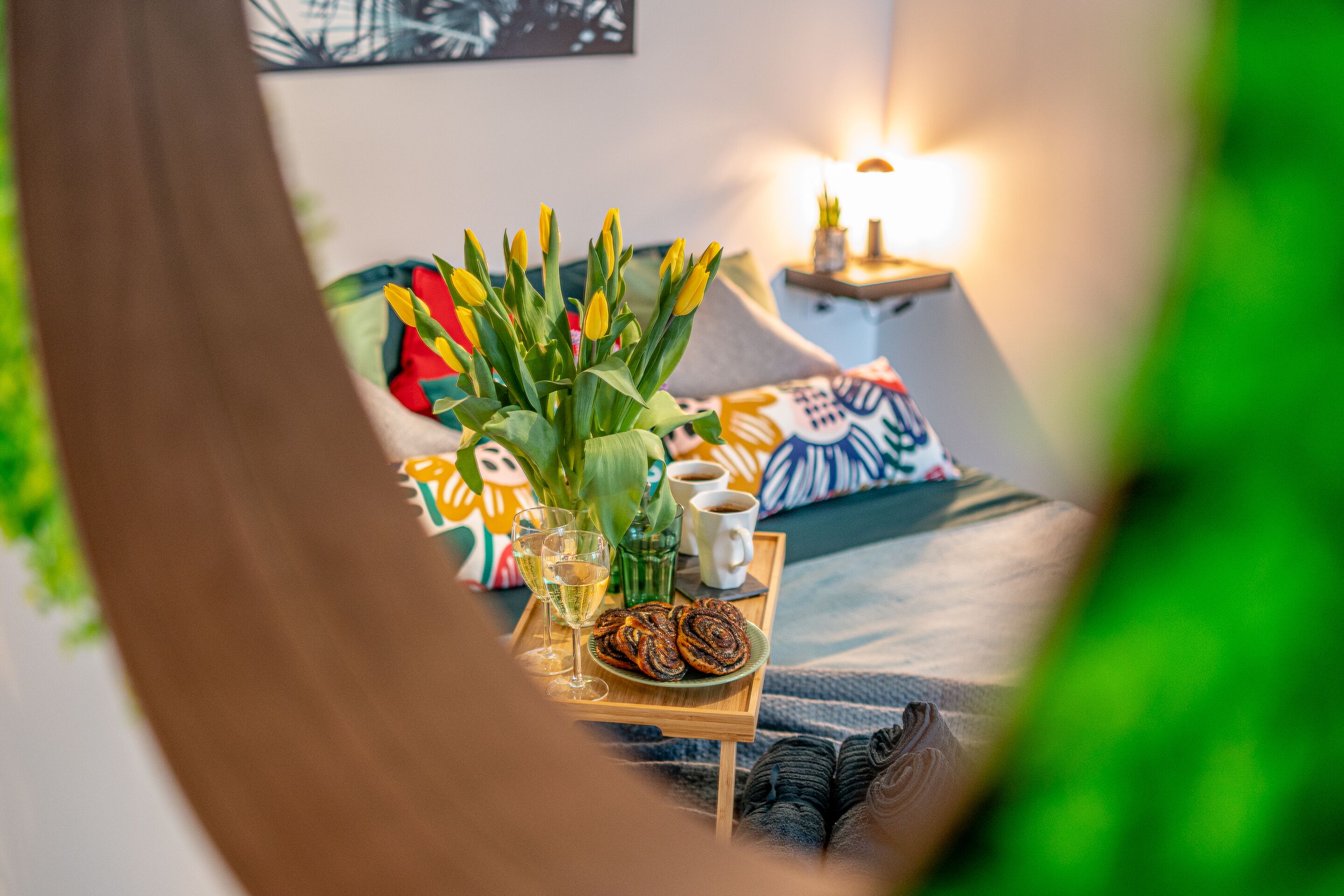 A cozy room with a bed, colorful pillows, a tray with pastries, glasses, and a vase of flowers.