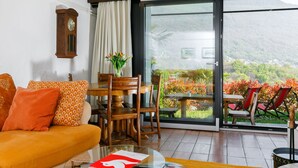 A living room with a sofa, a dining table with chairs, a coffee table, a clock, and a view of a garden through a sliding glass door.