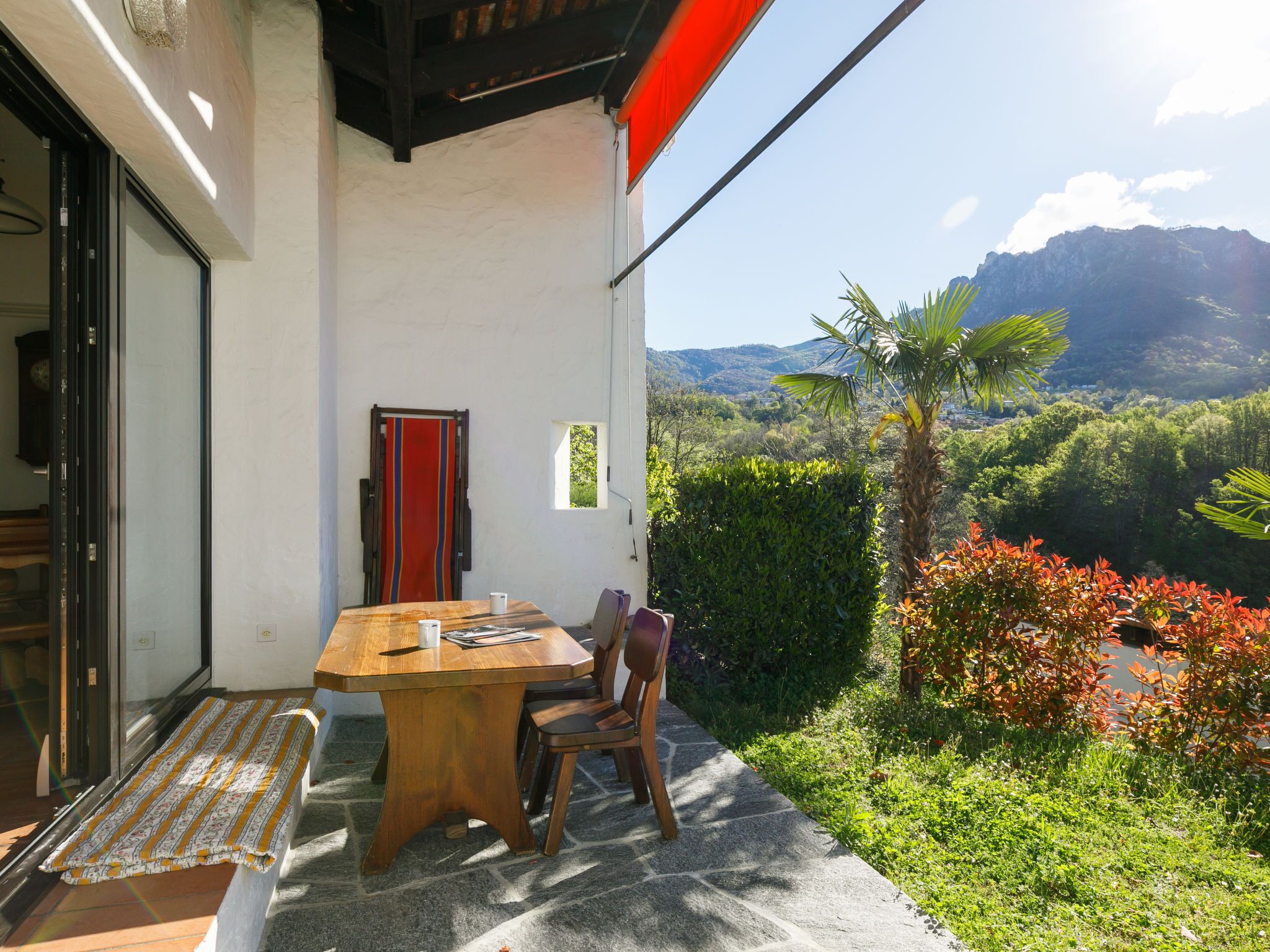 Une table en bois avec des chaises, placée à l’extérieur d’un bâtiment, une porte rouge et une vue sur des collines et des montagnes verdoyantes.
