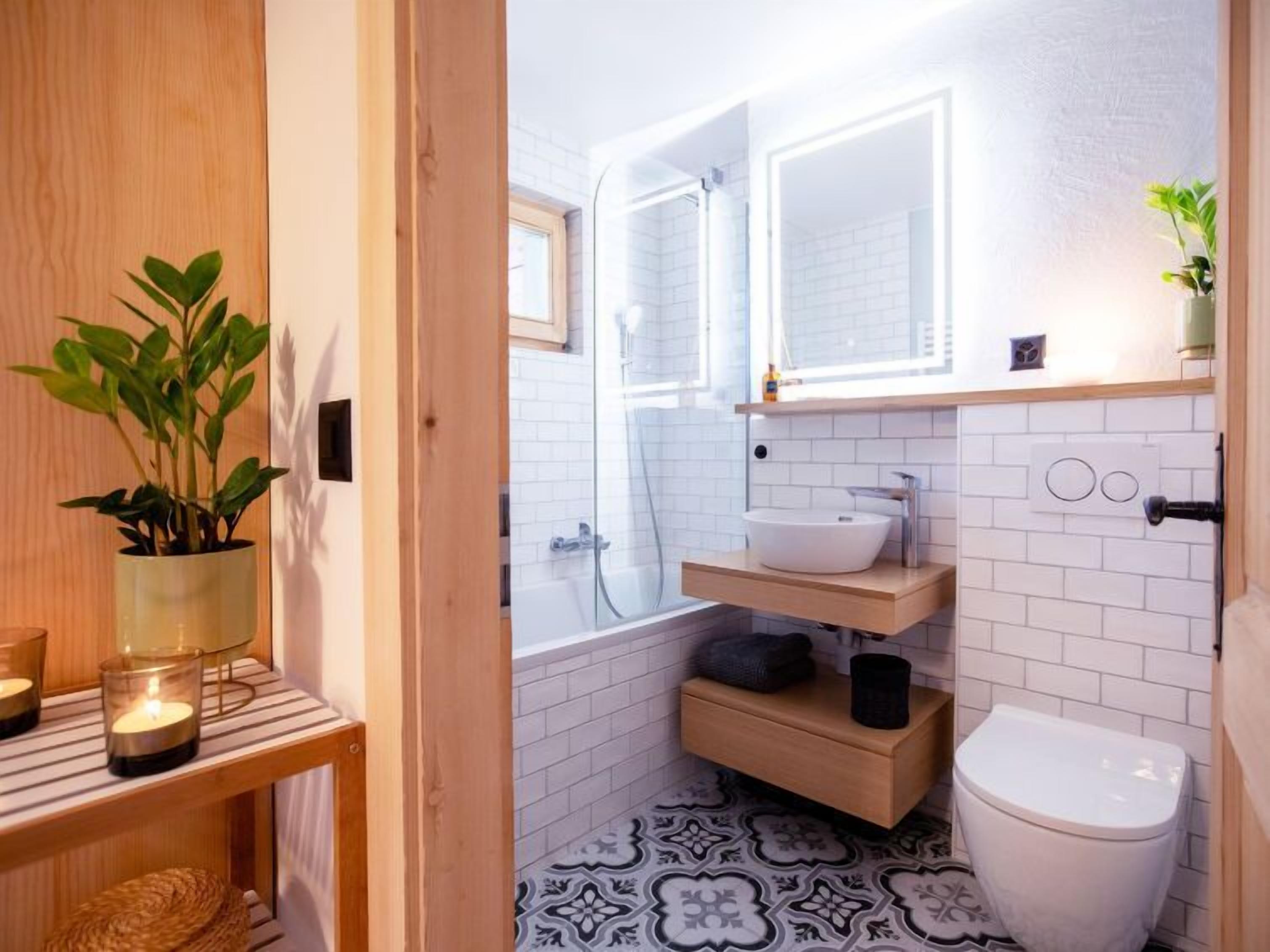 Une petite salle de bain avec un sol carrelé de blanc, des toilettes blanches, un lavabo avec un miroir au-dessus, et une étagère ornée d’une plante et de bougies.