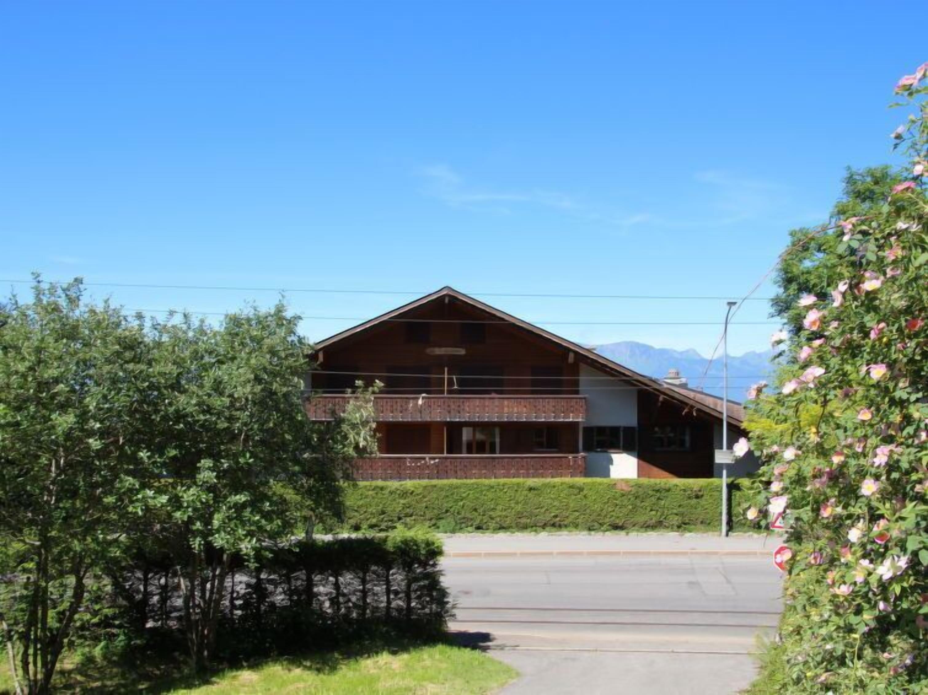 A two-story wooden building with a balcony, surrounded by greenery and a clear sky.