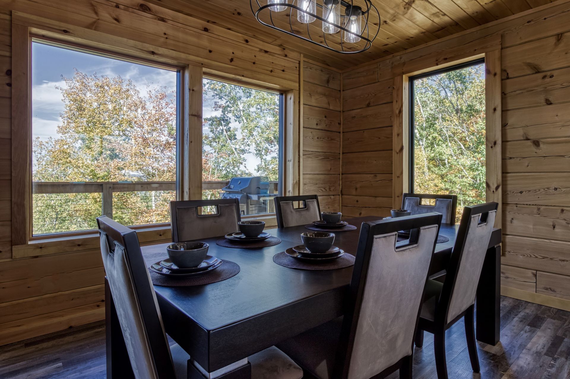 Comedor de madera con una mesa dispuesta para cuatro, ventanas amplias y una lámpara colgante.