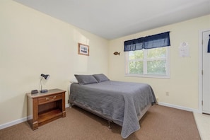 Quarto com cama, mesa de cabeceira, janela com persianas e um enfeite de peixe na parede.