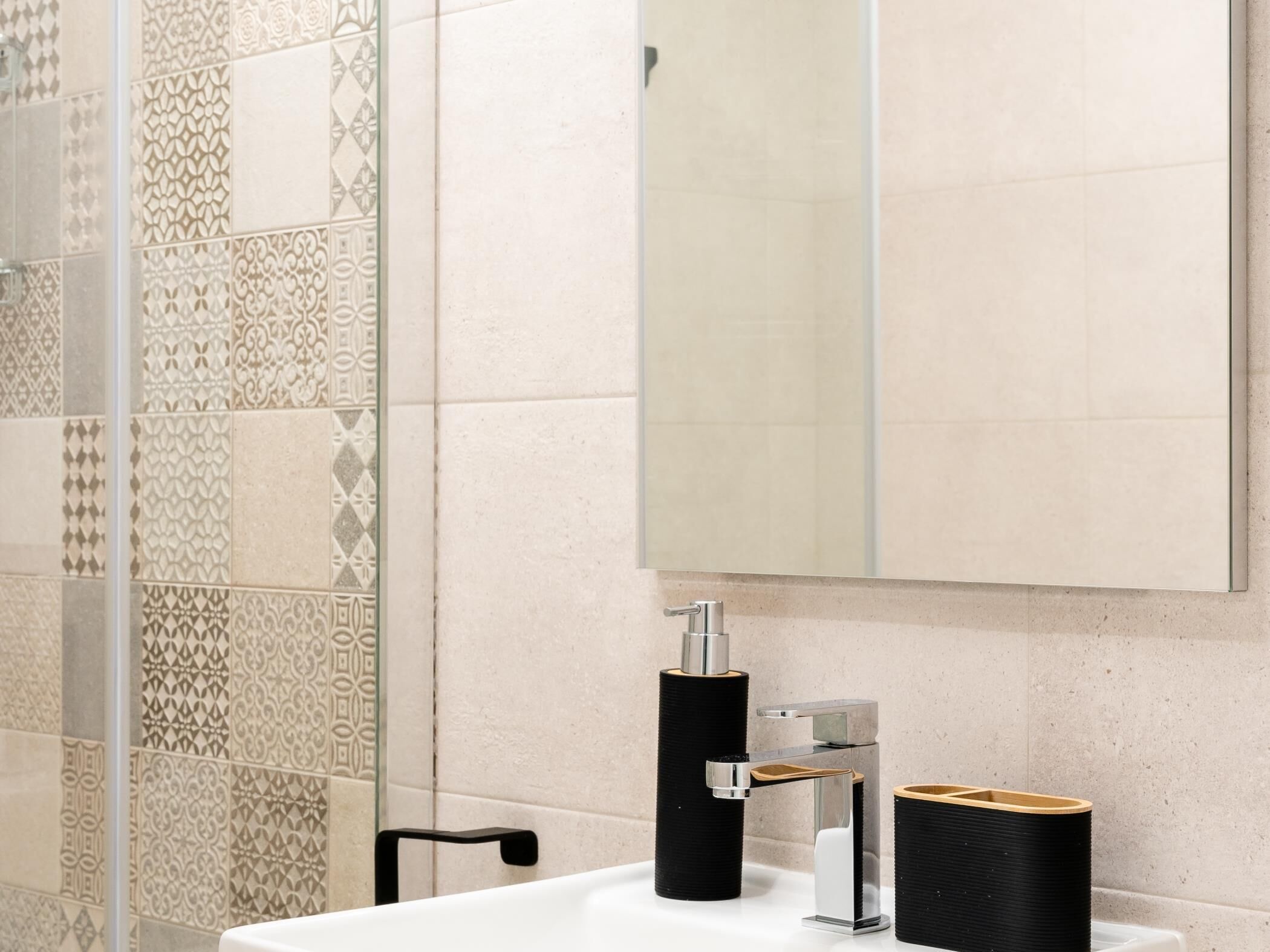 A bathroom with patterned tiles, a white sink, a mirror, and a black towel dispenser.